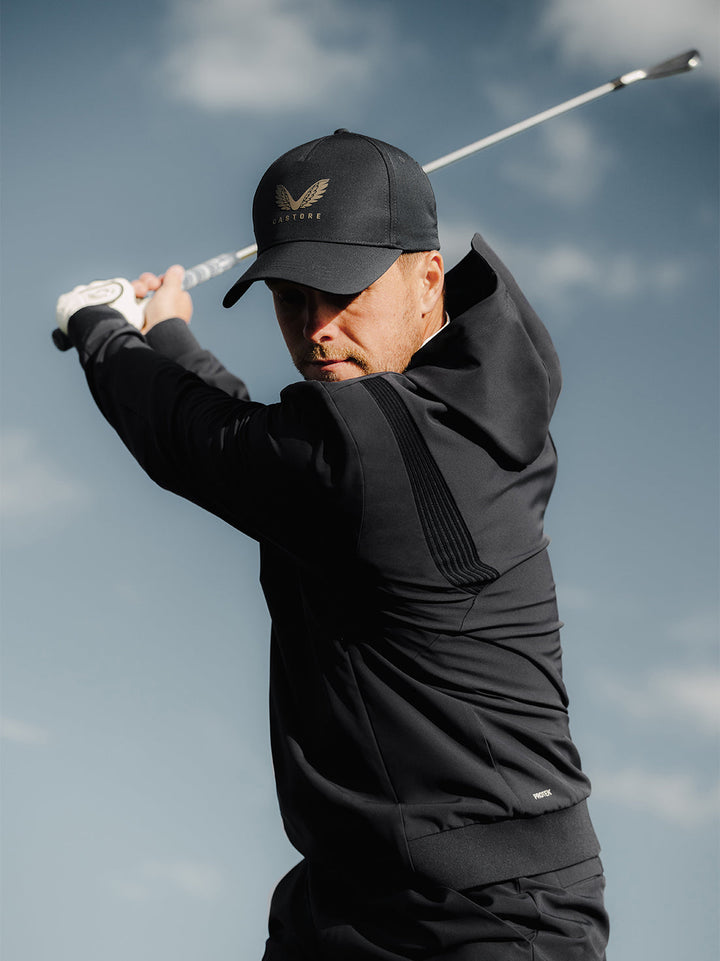 Golfer wearing black Pinnacle Performance Hoodie and cap mid swing against a blue sky.