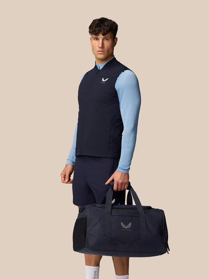 Male model wearing navy Apex Training Gilet over a light blue top, carrying a navy gym bag