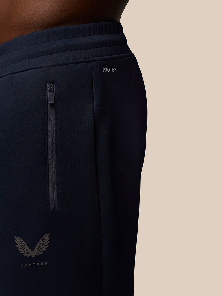 Close-up of navy joggers showing a vertical back zipper pocket, Protek branding at the waistband, and Castore logo at lower left.