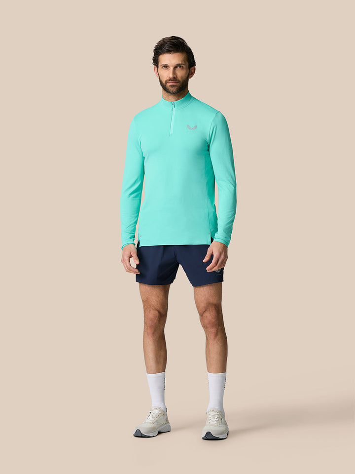 Male model wearing light aqua Zone Training 1/4 Zip Top with navy shorts standing against a beige backdrop.