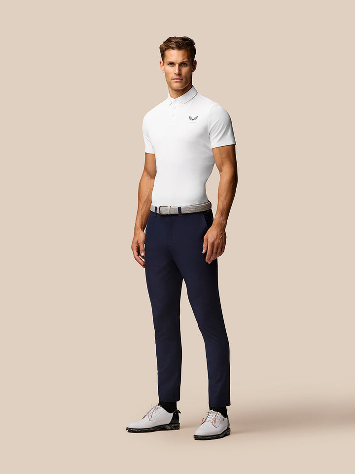 Front view of a man in a white Golf Club Essential Polo with Castore logo on the left chest, paired with navy trousers and a woven belt