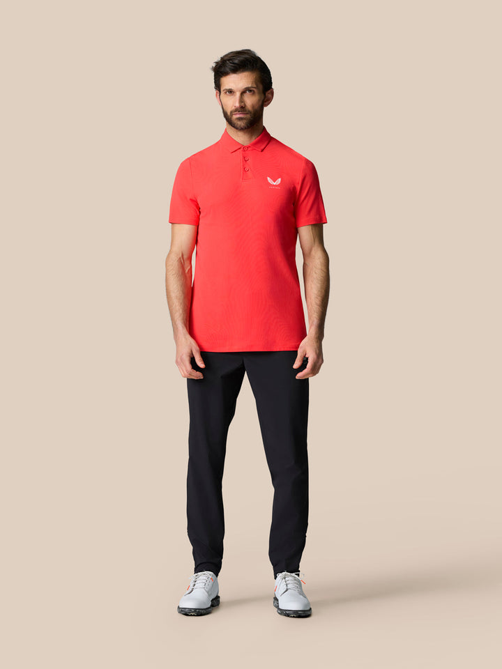 Full-body image of a man in a red polo with white Castore logo, paired with navy pants and white golf shoes.