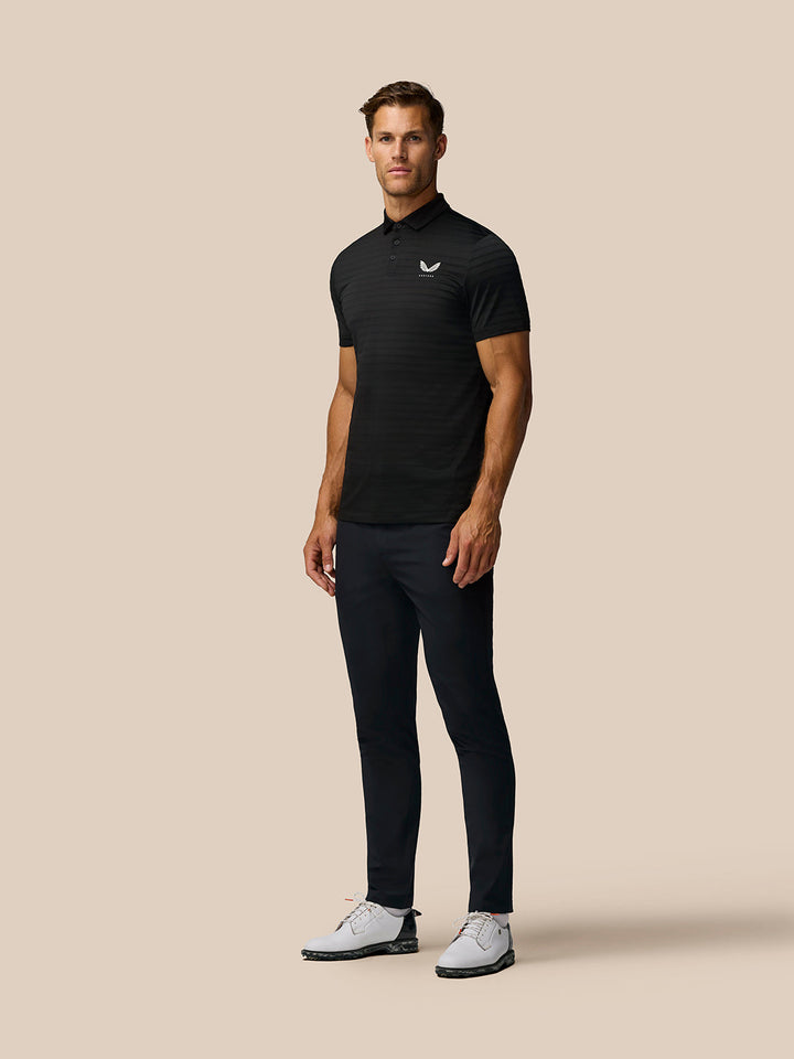 Male model in black Pinnacle Pique Stripe Polo with striped pattern left chest logo paired with dark trousers