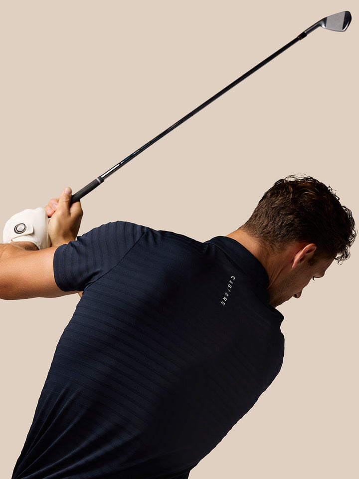 Golfer in navy Pinnacle Pique Stripe Polo mid swing against a beige background.