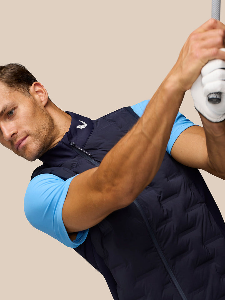 Male golfer wearing navy Pinnacle Gilet over a light blue shirt, gripping a golf club overhead.