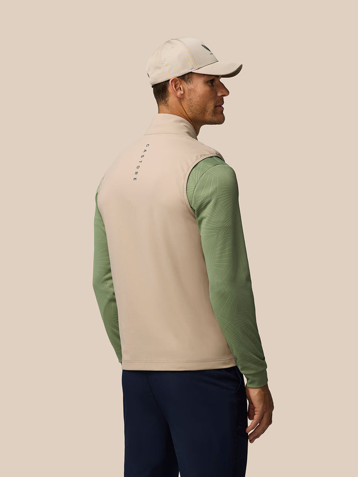 Back view of a man wearing a beige Golf Pinnacle gilet over green sleeves, CASTORE lettering runs down the back against a neutral backdrop.