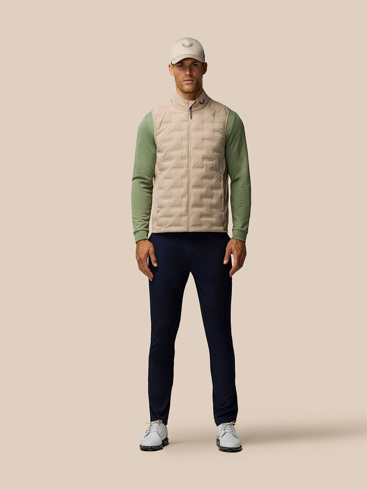 Full body front view of a man wearing a beige Pinnacle gilet with green sleeves, navy trousers and cap ready for a round of golf.