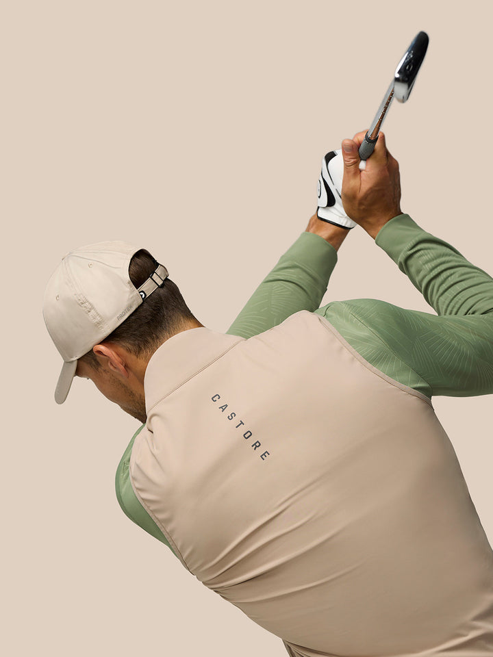 Golfer in beige Pinnacle gilet with green sleeves mid swing, captured from behind with cap and glove in neutral backdrop.