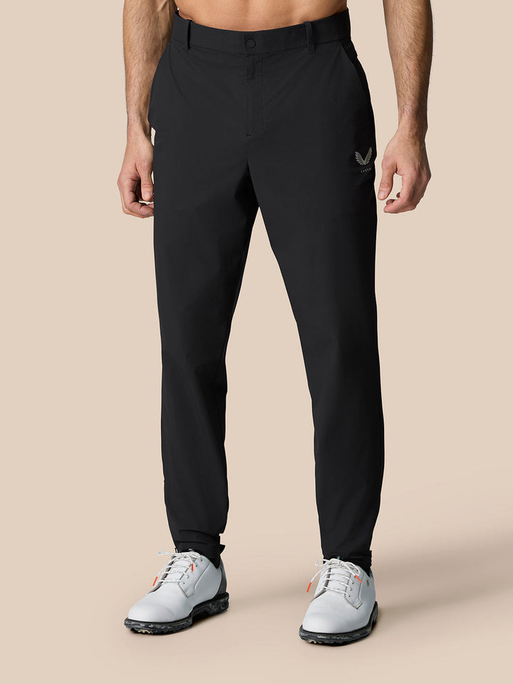 Full-length shot of a male model wearing black Golf Pinnacle 32 trousers with an orange polo, Castore branding on the right thigh.