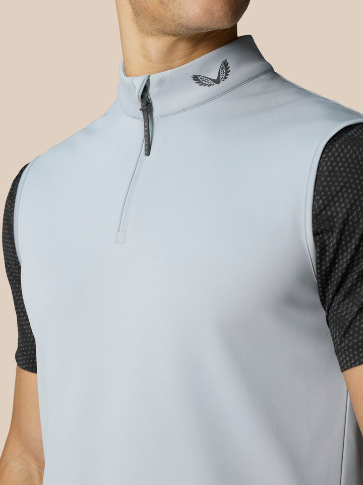Close up of light grey golf gilet front showing half zip and collar logo against a neutral beige background.