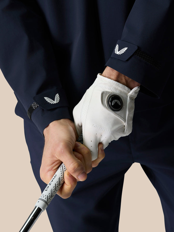 Close-up of a golfer gripping a club in a navy LVS Woven Jacket with wing emblem on the cuff and vent mesh