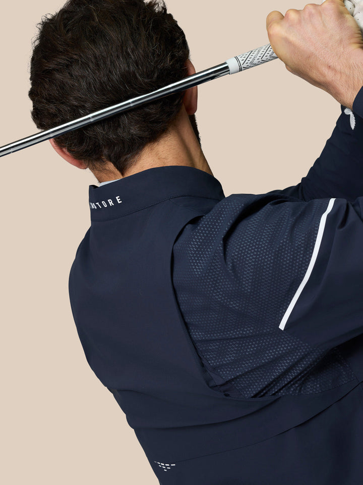 Back view of a navy Golf Pinnacle LVS Woven Jacket during a golf swing showing collar branding and vent panels.