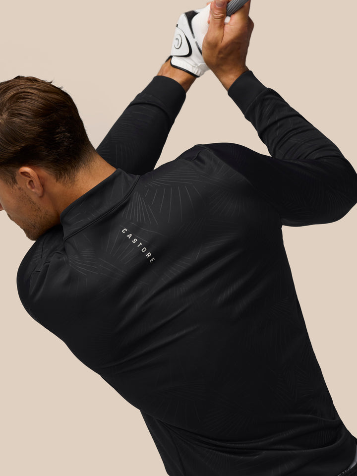 Close up of a man in a black quarter zip golf top with front zipper and all over print; Castore wing logo on chest.