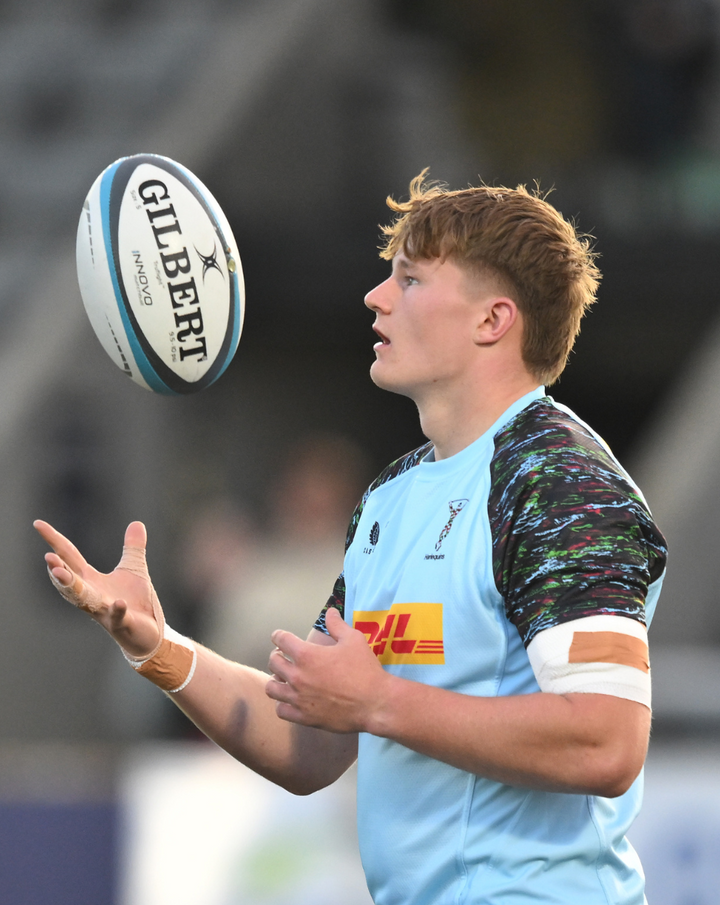 Male rugby player wearing sky blue Harlequins training tee with multicolored sleeves and a Gilbert rugby ball in the air